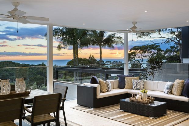 A bright, open-plan living and dining area with expansive floor-to-ceiling glazing overlooking a coastal ocean view at sunset. The space features a large L-shaped outdoor-style sectional, patterned cushions, a low coffee table, a dining table and chairs, ceiling fans, pale tiled flooring, and a glass balustrade extending to an elevated terrace.