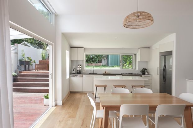 Bright, open-plan kitchen and dining area with white cabinetry, a long central island, timber flooring, and a large timber dining table with white chairs. Full-height glazing opens to a timber deck and landscaped outdoor area, while a woven pendant light adds a natural accent.