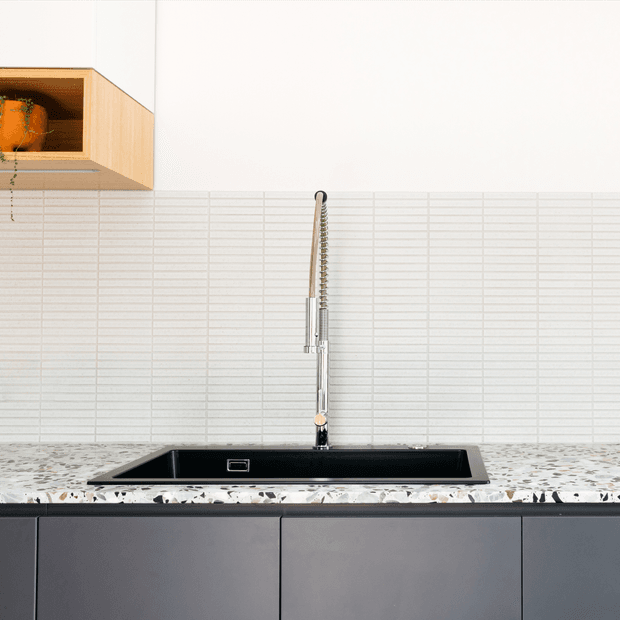 Close-up view of a contemporary kitchen sink area featuring a black undermount sink, tall chrome pull-out mixer tap, white horizontally ribbed tiled splashback, terrazzo-style benchtop, and matte dark grey cabinetry. A light timber open shelf with a plant is visible at upper left.