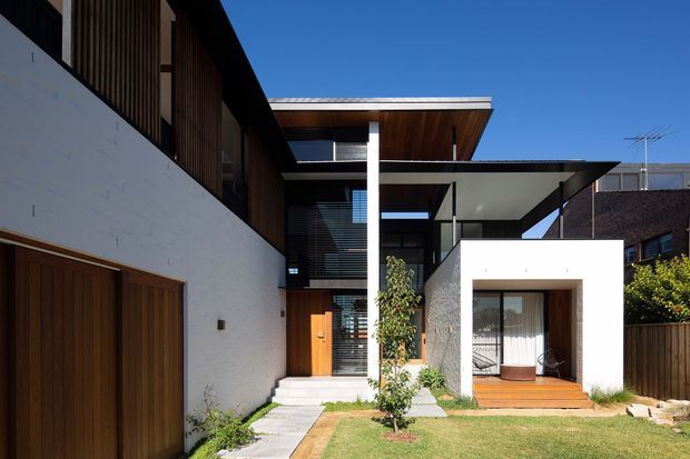 Contemporary multi-level residence with crisp white masonry walls, warm vertical timber cladding, broad roof overhangs, and extensive dark-framed glazing. The facade opens onto a small landscaped front garden with concrete stepping slabs, timber garage doors, and a sheltered timber entry deck. Strong geometric forms and shaded outdoor thresholds define the composition.