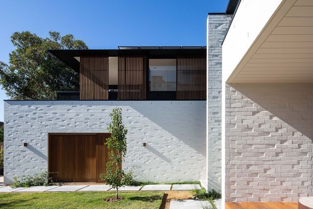 Exterior view of a contemporary two-storey residence with white painted brick walls, a dark flat roof, timber vertical screening on the upper level, and a large timber entry door. Strong geometric forms, deep overhangs, and recessed outdoor space create a crisp modern facade.