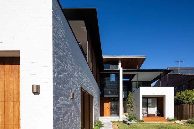A contemporary multi-level residential facade viewed from the front courtyard. The composition features white rendered masonry, a dark textured stone or brick side wall, warm timber doors and soffits, projecting flat roof planes, deep eaves, black-framed glazing, and a small landscaped entry path. Strong geometric forms contrast with natural timber and greenery under a clear blue sky.