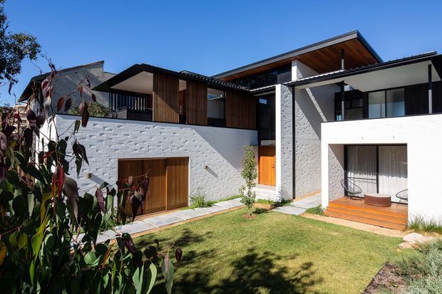 Exterior view of a contemporary multi-level residence arranged around a landscaped front courtyard. The house combines white textured masonry walls, dark metal-framed glazing, warm timber cladding and soffits, flat and low-pitched roof forms, and a covered timber-decked outdoor sitting area. A timber garage or entry door is set into the masonry wall, with balconies and recessed openings above.