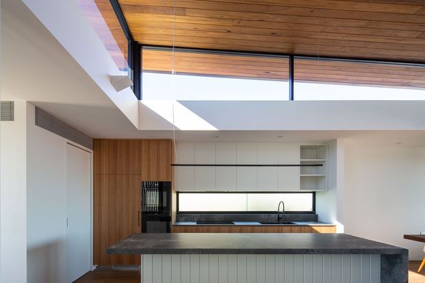 A bright contemporary kitchen with a large central island, white flat-panel cabinetry, warm timber wall cladding and ceiling lining, dark stone island benchtop, and a long horizontal window above the sink. High-level glazing and a partially double-height ceiling bring in abundant natural light.