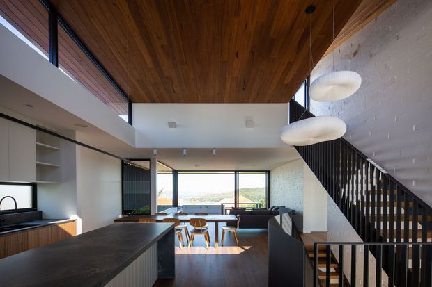 Open-plan contemporary interior showing a kitchen in the foreground flowing into a dining and living area. The space features a dark stone kitchen island, white fixed cabinetry, extensive glazing, a white-painted brick wall, a timber-lined ceiling, dark timber flooring, and a black open-riser staircase with sculptural pendant lights. A distant landscape is visible through the windows.
