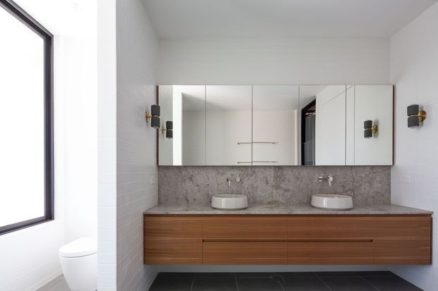 Contemporary bathroom vanity with a long floating timber cabinet, two white vessel basins, a grey stone-look benchtop and matching textured splashback. A wall-to-wall mirrored cabinet sits above the vanity, with black and brass wall sconces, white tiled walls, dark floor tiles, and a partially visible toilet beside a large window.