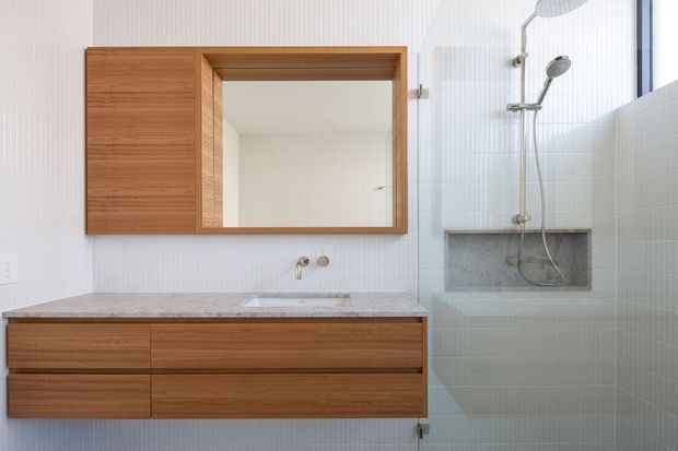 Bright contemporary bathroom with a wall-mounted timber vanity featuring wide drawers, a pale stone benchtop and integrated basin, and a matching timber-framed mirrored cabinet. The shower area has vertical white wall tiles, a frameless glass screen, chrome fittings, and a recessed grey stone niche. The room has a clean, minimal palette of white, light timber, pale grey and chrome.