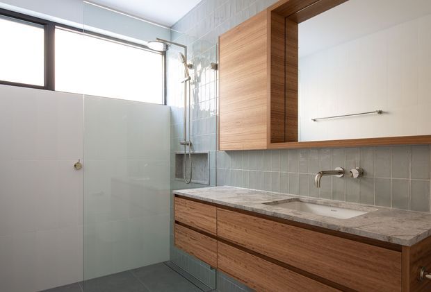 Bright contemporary bathroom featuring a long timber vanity with integrated basin, grey stone benchtop, grey-blue tiled splashback and walls, frameless glass shower screen, wall-mounted chrome tapware, and a large timber-framed mirrored cabinet. A high horizontal window provides natural light.