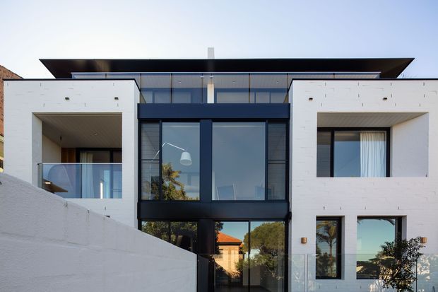 Front exterior view of a contemporary multi-level residence with a strong geometric composition. White rendered masonry volumes frame a dark charcoal glazed central section, with extensive floor-to-ceiling glazing, recessed balconies, a projecting low-profile roof, and a partially concealed entry. A white boundary wall occupies the foreground.