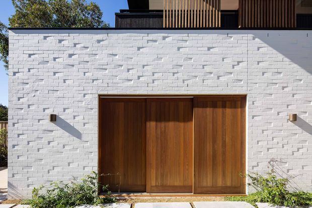 Exterior architectural detail of a contemporary residence, featuring a white-painted textured brick wall, large warm timber sliding or folding doors, minimalist wall lights, and a dark upper-level balcony with vertical timber screening. Landscaping is visible along the base.