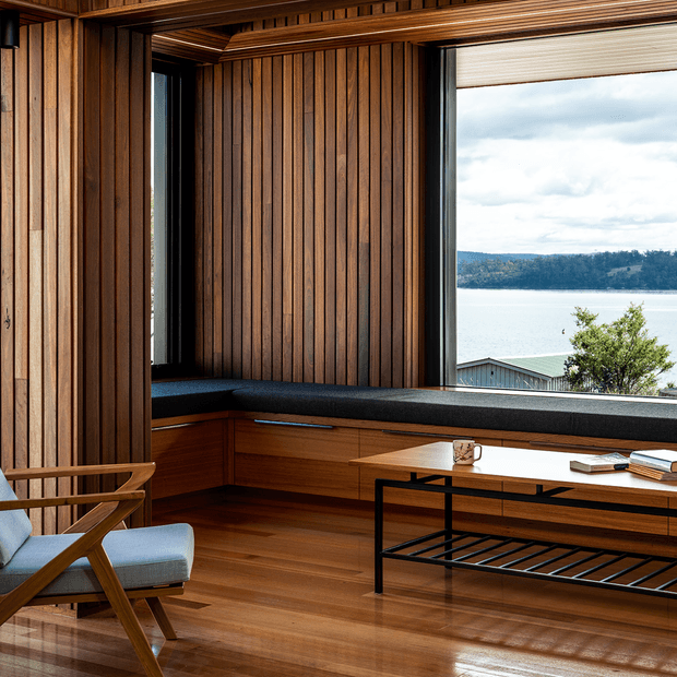 A warm, wood-lined contemporary living room or study with extensive vertical timber wall panelling, a built-in window seat, large black-framed windows, polished timber flooring, and a minimalist black metal coffee table. A timber lounge chair with pale blue upholstery sits in the foreground. The windows overlook a calm body of water and a wooded shoreline.