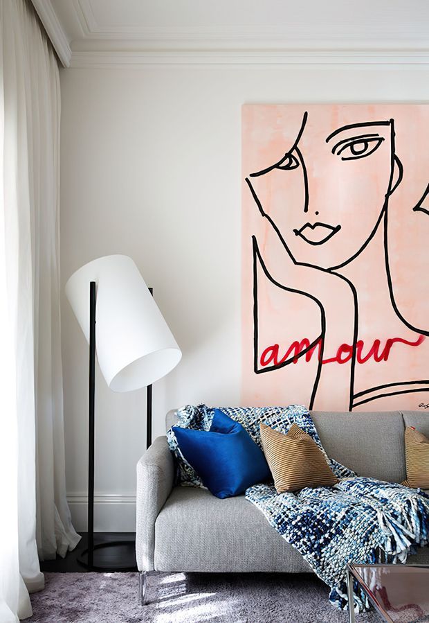 A refined living room with pale walls, decorative ceiling moulding, full-height sheer curtains, a sculptural white-and-black floor lamp, and a light grey sofa. The sofa is styled with a vivid blue cushion, striped beige cushion, and blue patterned throw. A large pink graphic artwork with a black line-drawn face and red lettering forms the main feature wall. A reflective glass-topped coffee table and plush grey rug complete the setting.