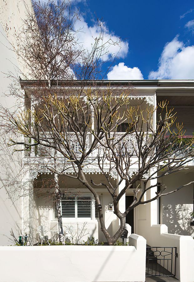 Front exterior view of a two-storey heritage-style residence with a pale rendered facade, covered verandah, ornate white decorative lacework, timber columns, and a low boundary wall with metal gate. A leafless pruned tree occupies the foreground, with strong sunlight and blue sky behind the building.