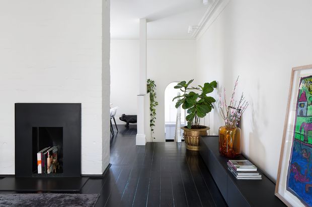 Bright, narrow contemporary interior with white walls, a black fireplace surround, and dark timber flooring. A long dark built-in console runs along the right wall, styled with potted plants, books, decorative vases, and framed artwork. An opening reveals a partially visible adjoining living area, while ceiling cornices and simple white columns add architectural definition.