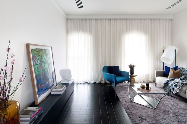 Bright, refined contemporary living room with floor-to-ceiling sheer curtains, dark timber flooring, a grey rug, sculptural coffee tables, a deep blue armchair, and a light upholstered sofa. A low dark built-in console displays artwork and decor, while a large angled floor lamp and floral branches add visual interest. The room has a high, detailed ceiling with linear air-conditioning grilles and abundant diffused daylight.