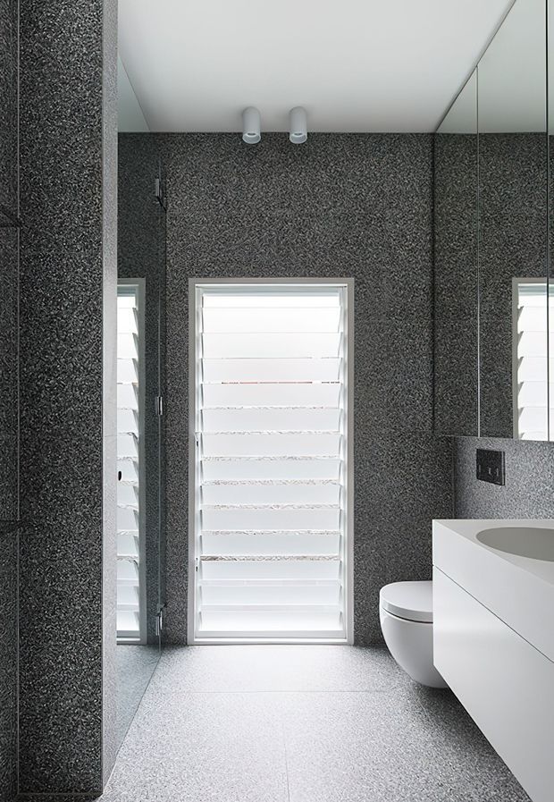 A compact contemporary bathroom with dark charcoal textured wall finishes, pale speckled flooring, a white wall-hung vanity, integrated basin, wall-mounted toilet, large mirror, and white plantation shutters. The space has clean geometric lines and bright natural light.