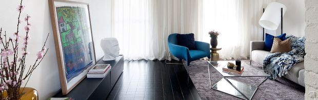 A bright, contemporary living room with dark timber flooring, sheer white curtains, a teal-blue accent armchair, a neutral sofa with patterned textiles, and a geometric glass coffee table. A large framed artwork leans against the wall beside a low dark console, while a tall white floor lamp and floral branches add decorative detail.