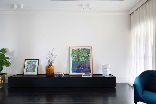 Bright, refined interior vignette featuring a long black low-profile console against a white wall, colourful framed artwork, a glass vase with branches, and a sculptural white object. Dark timber flooring, full-height sheer curtains, recessed ceiling detailing, and a deep blue upholstered armchair are visible. The composition has a clean contemporary feel with strong light-dark contrast.
