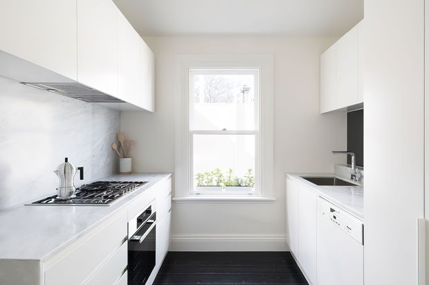 Bright, compact white galley kitchen with full-height white cabinetry on both sides, pale marble-look splashback and benchtops, integrated stainless-steel appliances, a gas cooktop, sink, and a central window. Dark timber flooring creates contrast against the predominantly white fit-out.