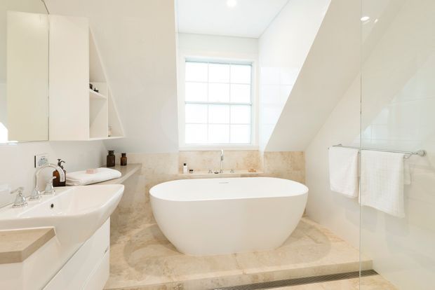 Bright, refined bathroom with a freestanding white oval bathtub positioned beneath a window, a wall-mounted vanity with basin and mirrored cabinetry, and a frameless glass shower enclosure. Pale marble-look stone surfaces and white finishes create a clean, airy appearance. The sloping ceiling suggests an attic or upper-level bathroom.