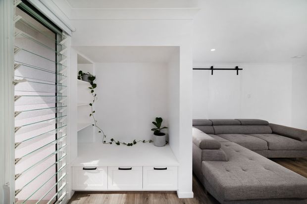 Bright minimalist living room with a large grey modular sectional sofa, white built-in cabinetry and open shelving, a trailing indoor plant, timber flooring, and a black barn-style sliding door rail. Plantation-style louvred window panels provide daylight from the left.
