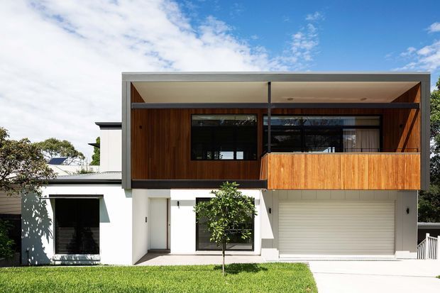 Front exterior view of a contemporary two-storey house with a strong rectangular composition, white rendered lower level, warm vertical timber cladding across the upper facade and balcony, dark-framed glazing, a projecting flat-roofed upper volume, and an integrated single garage. A small lawn and young tree sit in front of the home under a bright blue sky.