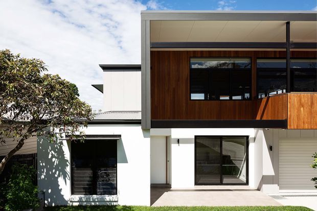 Modern multi-level residential exterior with a crisp white rendered facade, dark metal framing, extensive glazing, and a prominent upper-storey section clad in warm vertical timber. The design includes projecting box forms, a covered ground-floor terrace or entry zone, and a partially visible timber balcony screen. Landscaping is limited to a tree at the left edge.