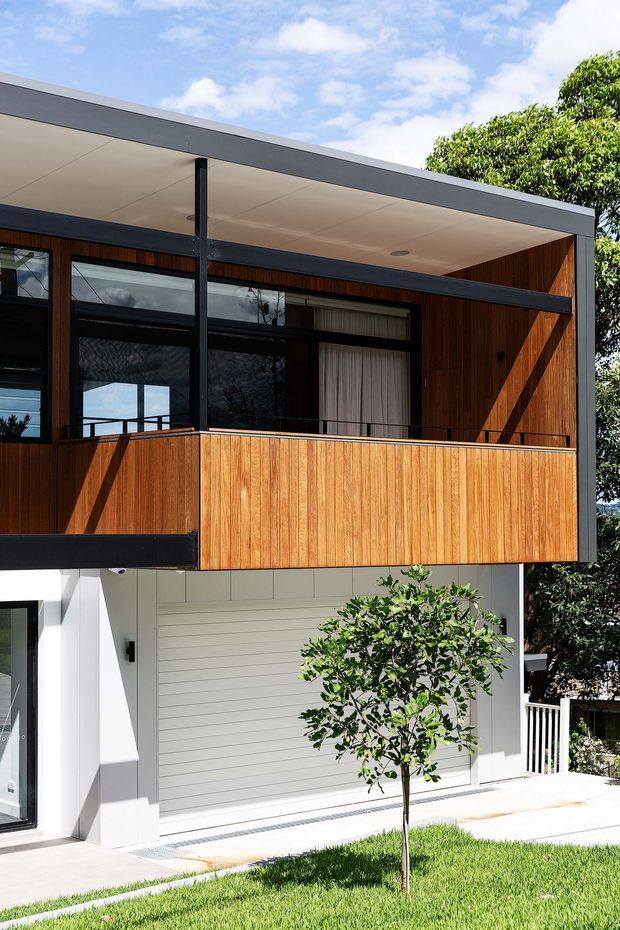 Exterior view of a contemporary two-storey residence with a flat roof, dark metal framing, warm vertical timber cladding, broad glazed openings, and a projecting upper-level balcony screened with timber panels. The lower level has white horizontal cladding and a garage door, with a small landscaped lawn and tree in front.