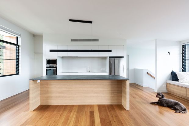 Bright open-plan kitchen with a large timber island, dark stone benchtop, integrated stainless-steel appliances, white cabinetry and splashback, and timber flooring. A bedroom area is visible to the right, with a dog resting on the floor. Contemporary detailing includes streamlined black pendant lighting, white walls, timber-framed joinery, and a stair opening.