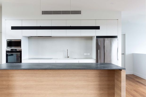 Bright contemporary kitchen with a large timber-fronted island, white full-height cabinetry, integrated stainless-steel appliances, a pale splashback, and a dark stone-look benchtop. The clean linear detailing and concealed storage create a minimalist, refined appearance.