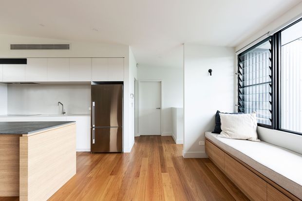 Bright open-plan interior combining a minimalist white kitchen with a timber island, stainless-steel refrigerator, warm timber flooring, and a built-in window seat with storage. Large louvred windows bring in natural light, while a short hallway and closed internal door are visible beyond the kitchen.