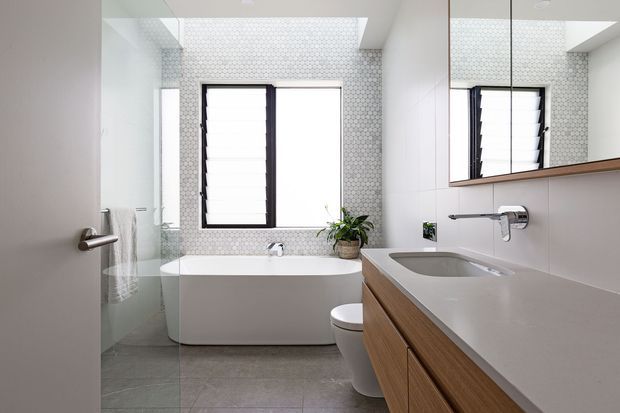 Bright contemporary bathroom featuring a freestanding white bathtub beneath a large window, a wall-mounted timber vanity with integrated basin, mirrored cabinetry, a wall-hung toilet, and pale patterned mosaic wall tiles. A glass shower enclosure is visible at the left.