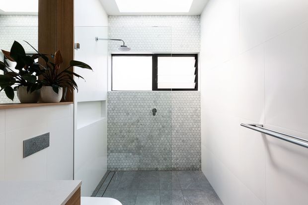 Bright contemporary bathroom with a walk-in shower, white wall surfaces, grey hexagonal mosaic tile shower walls, large grey floor tiles, black-framed awning window, linear chrome towel rail, skylight, recessed shelving, and built-in white vanity cabinetry with indoor plants.