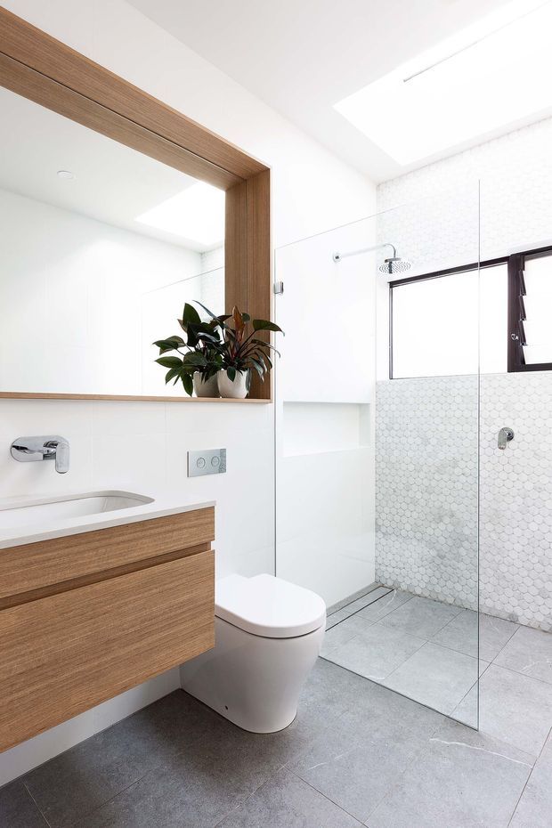 Bright contemporary bathroom with a floating timber vanity, integrated white basin, large timber-framed mirror, wall-hung toilet, frameless glass shower enclosure, and white hexagonal wall tiles. Large grey floor tiles and a skylight contribute to the clean, light-filled appearance.