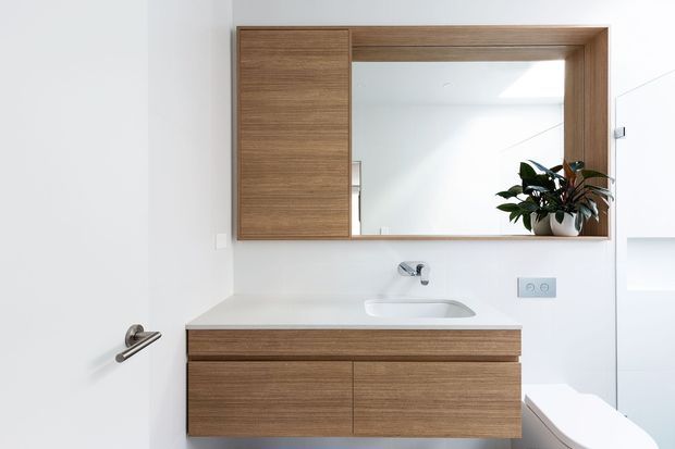 Bright, minimalist bathroom vanity area with a wall-mounted warm timber bathroom cabinet and integrated mirrored storage above. The white basin, chrome tapware, white walls, and partially visible freestanding bathtub create a clean, contemporary fit-out. A potted plant adds a small natural accent.