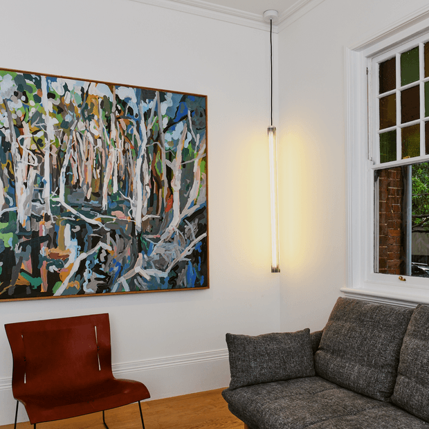 A refined living room corner with white walls, detailed traditional trim, a large colourful abstract artwork, a slim hanging wall light, a grey textured sofa, a rust-toned leather chair, timber flooring, and a multi-pane window overlooking greenery and brickwork.