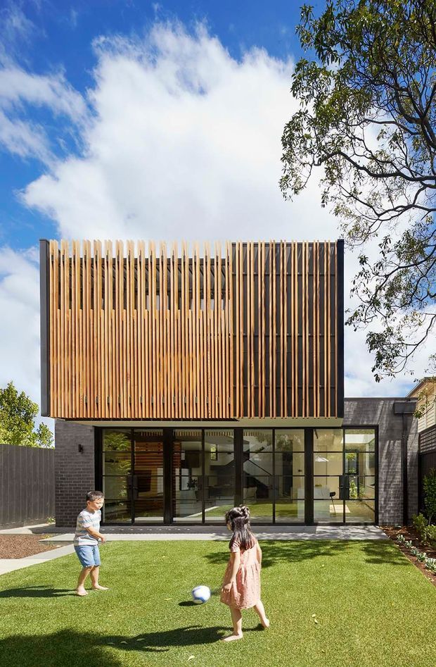 A contemporary two-storey residential facade viewed from the rear garden, featuring a large upper-level screen of closely spaced vertical timber battens, dark masonry side walls, and extensive black-framed ground-floor glazing. A lawn and children are in the foreground, with mature trees and a bright sky surrounding the home.