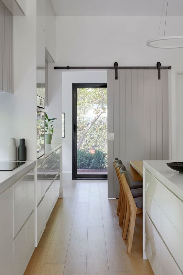 Bright, narrow contemporary kitchen interior with extensive white built-in cabinetry, pale timber flooring, a central white island with integrated seating, and a grey vertical-panel barn-style sliding door on black hardware. A black-framed glazed exterior door brings in garden views, while a slim circular pendant light and minimal decor reinforce the clean modern design.