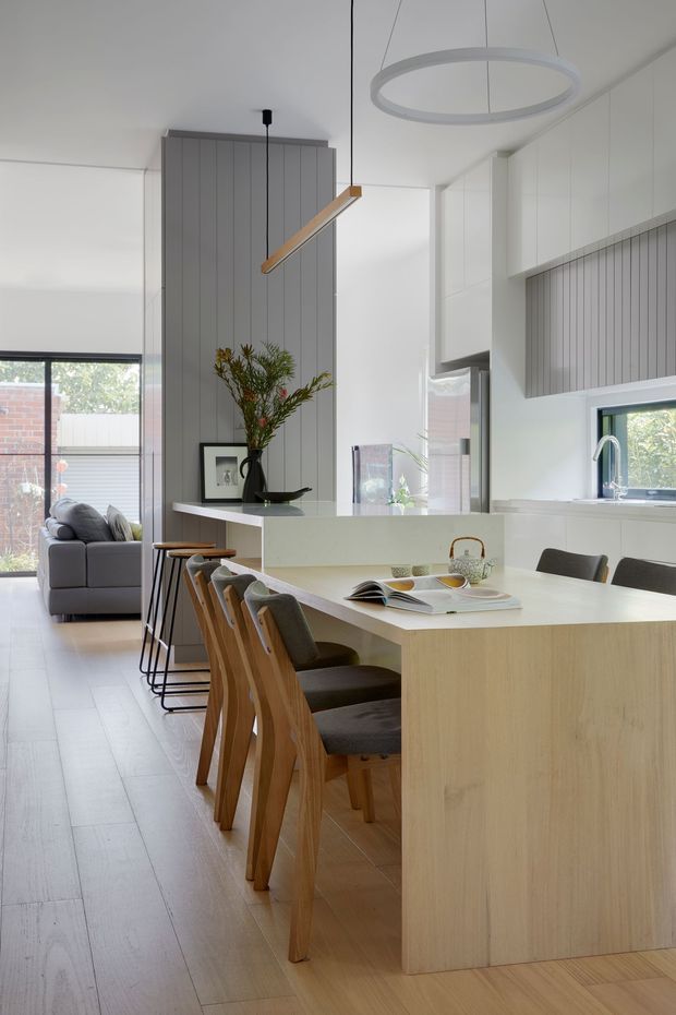 Bright open-plan kitchen and dining area with a large pale timber island, integrated white cabinetry, grey vertical-panel feature joinery, timber dining chairs, pendant and circular ceiling lights, and a living room visible beyond. The space has a clean, contemporary linear design with abundant natural light and light timber flooring.