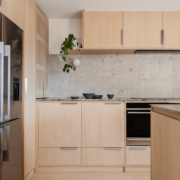 A warm, minimalist contemporary kitchen with pale timber cabinetry, a full-height pale stone tiled splashback, integrated black appliances, and a timber island in the foreground. A vertical timber slat partition and stainless-steel refrigerator frame the left side, while restrained black ceramic accessories and a hanging plant add contrast.