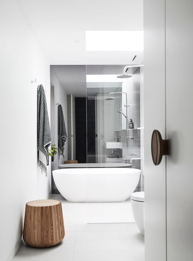 A bright, compact contemporary bathroom viewed through a partially open door. The space features a freestanding white bathtub, wall-mounted white basin, glass shower enclosure, large mirrored wall, white tiled floor and walls, chrome fixtures, and a timber stool. A black-and-white towel hangs on the wall, and the mirror creates repeated reflections of the bathroom.