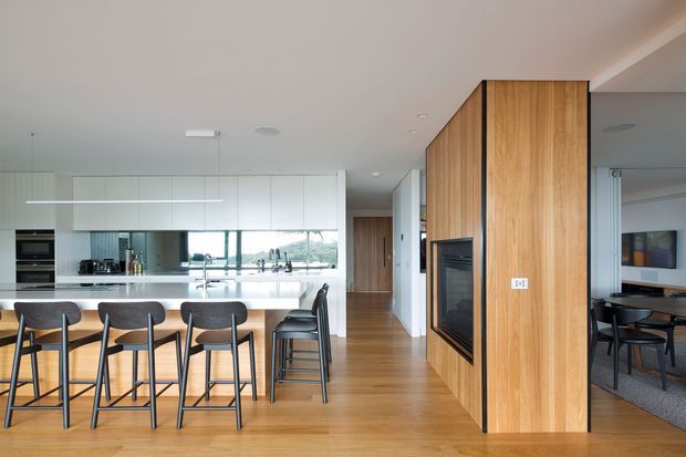 Bright open-plan contemporary interior featuring a kitchen with a long timber island and four black bar stools, white cabinetry, integrated appliances, and a mirrored backsplash reflecting the view outside. Warm timber flooring continues through the hallway, beside a full-height timber-clad fireplace wall. A dining area is visible through the opening on the right.