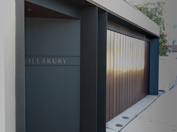 Contemporary dark-painted building entrance and garage frontage, featuring a large vertically slatted timber-toned garage door, deep recessed doorway, and the name “VILLARUBY” integrated into the wall panel. A narrow concrete pedestrian path runs alongside the facade.