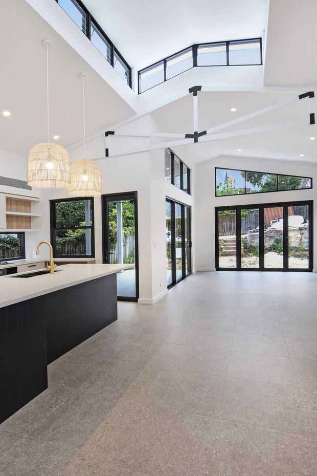 A bright, open-plan contemporary kitchen and living area with a dramatic double-height ceiling and mezzanine-level windows. The kitchen features a large dark island with a pale benchtop, integrated shelving, a brass-toned tap, and woven pendant lights. White walls and ceilings contrast with black-framed floor-to-ceiling glazing and clerestory windows, while the spacious grey tiled floor extends toward glazed exterior doors.