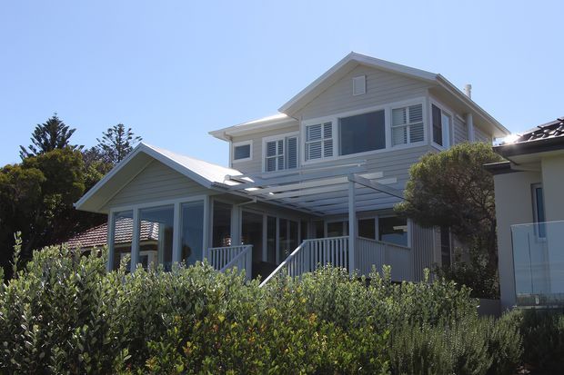 Exterior view of a light grey, two-storey weatherboard house with a pitched gable roof, white trim, shuttered windows, and a covered front veranda or outdoor living area. Dense hedging and established landscaping partially screen the lower facade.
