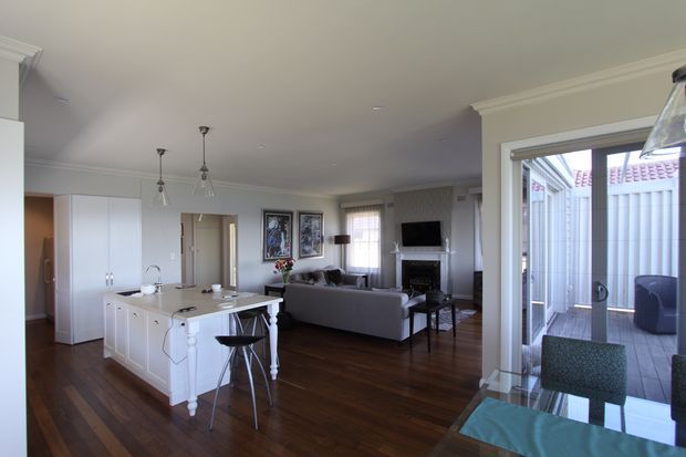 Open-plan residential interior combining a white kitchen with island, dining area, and living room. Dark timber flooring contrasts with white cabinetry and light walls, while French doors open to a small paved courtyard or outdoor area. Traditional detailing includes decorative island legs, pendant lights, framed wall art, and ceiling cornices.