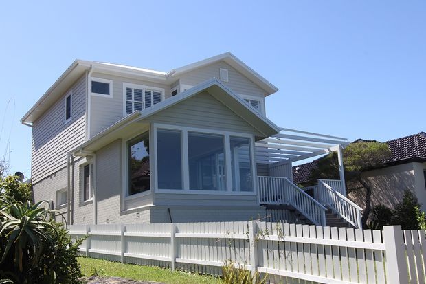 Exterior view of a two-storey light-coloured weatherboard house with multiple pitched gables, white-framed windows, a projecting enclosed front porch or sunroom, raised entry stairs, a pergola-like roof structure, and a white picket fence. The home sits in a suburban residential setting under clear blue sky.
