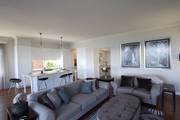 Bright open-plan living room with a grey sectional sofa and matching armchairs arranged around a large oval upholstered coffee table. The space includes a compact white kitchen with a peninsula and bar stools, pendant lights, dark timber flooring, white cabinetry, pale walls, framed artwork, and large windows providing natural light.
