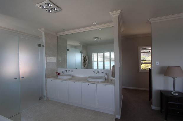 A spacious, elegant bathroom with a long white double vanity, integrated basins, large wall mirror, pale stone-look wall and floor finishes, frosted glass doors, decorative ceiling cornices, and recessed and multi-light ceiling fixtures. A bedroom or dressing area is visible through the opening on the right, with plantation shutters and a bedside table.