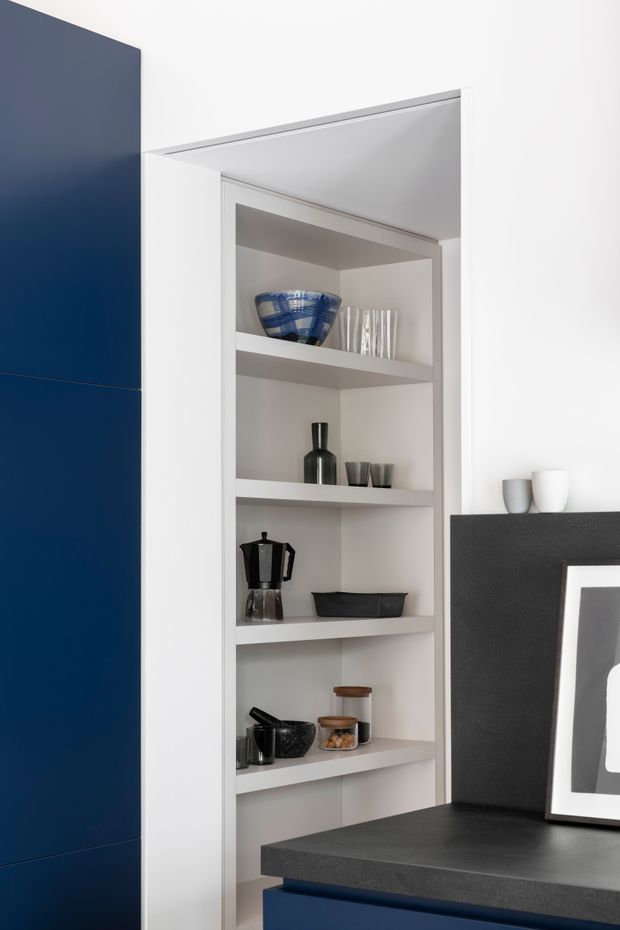 A close-up view of a contemporary built-in open shelving unit, likely forming part of a kitchen or dining area. The white recessed shelves display glassware, ceramics, a moka pot, bowls, and jars. Dark navy cabinetry appears at left and below, with a charcoal benchtop and a black-and-white framed artwork at right. The composition is clean, restrained, and high-contrast.
